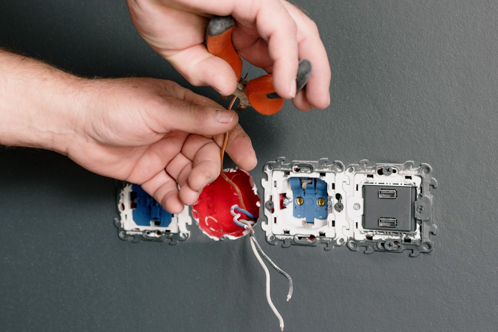 An electrician using pliers to terminate the wiring behind a wall socket during an outlet installation.